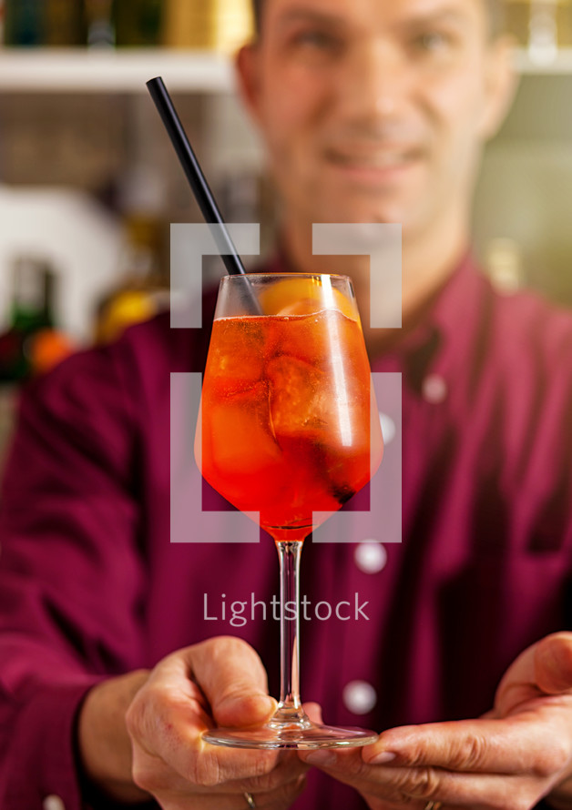 Smiling bartender in a vibrant purple shirt serving a refreshing aperol spritz at a bar