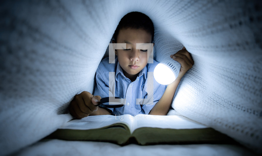 a little boy reading a Bible under a blanket holding a light