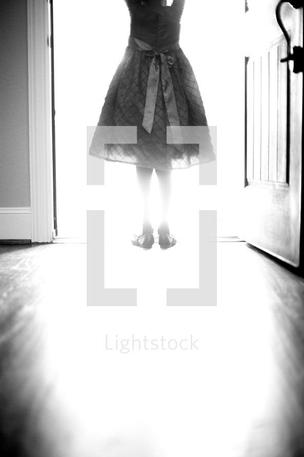 a girl child in a dress standing in light from an open doorway 