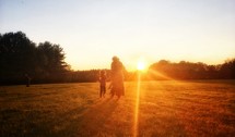 a mother and child walking outdoors through a field at sunset 