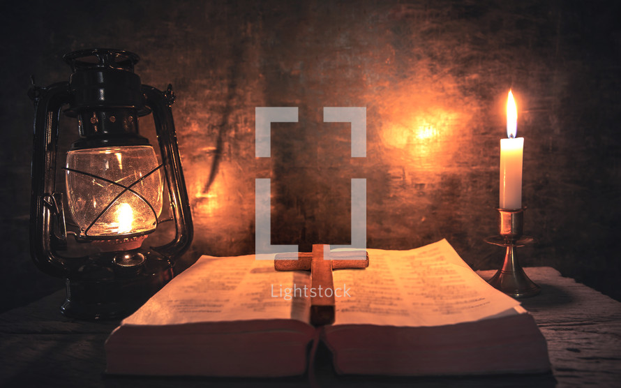 cross on an open Bible with light from a candle and oil lamp 