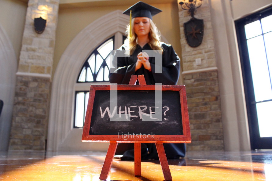 young woman in cap and gown praying on her knees in front of a sign that says where