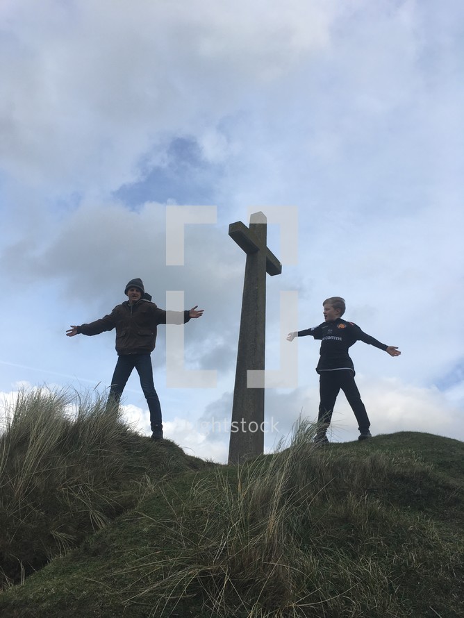 boy standing with open arms beside of a cross on a hilltop 