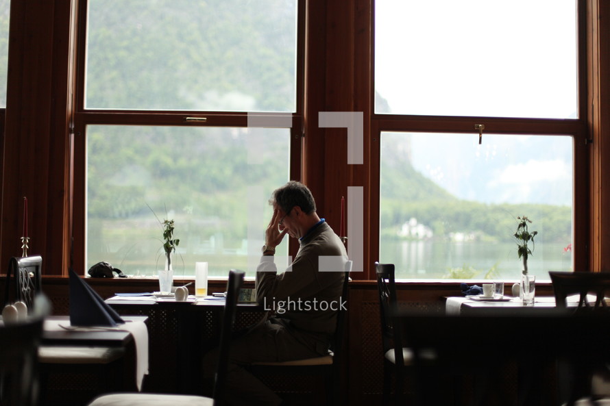 Man in prayer sitting at a table in front of a window in a restaurant.