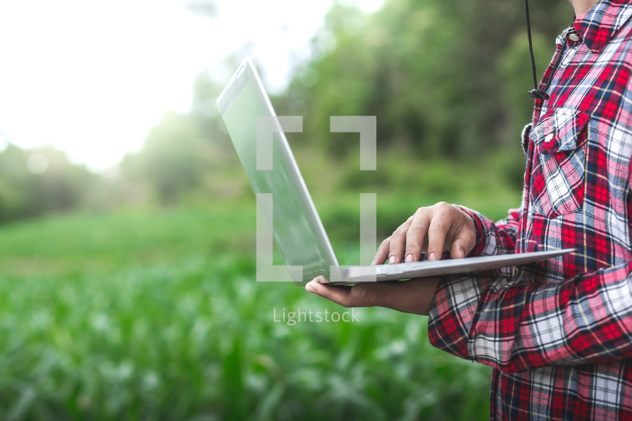 young farmer sitting and checking his corn field and working on laptop computer under the tree,