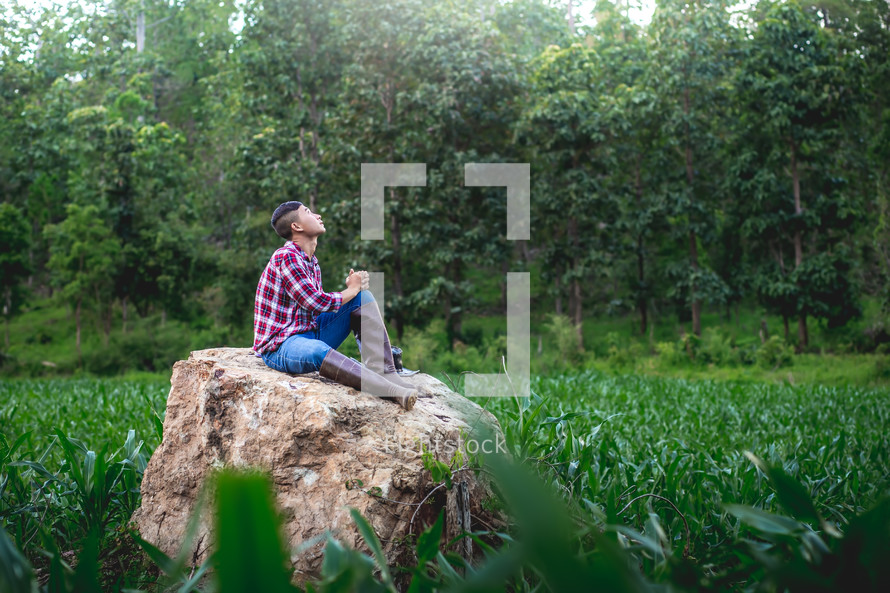 Young farmer sitting on a big stone praying over his corn field 