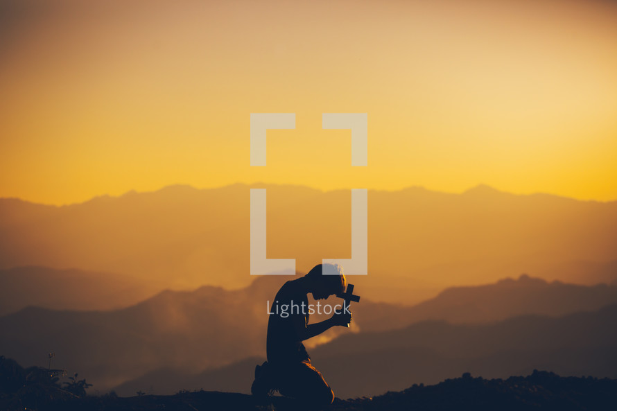 silhouette of a boy kneeling in prayer on a mountaintop at sunset 