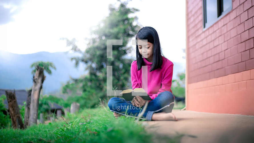 a girl in a pink jacket outdoors reading a Bible 