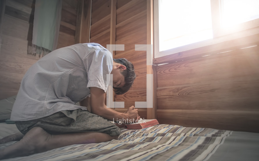 a teen kneeling on a bed by a window praying 
