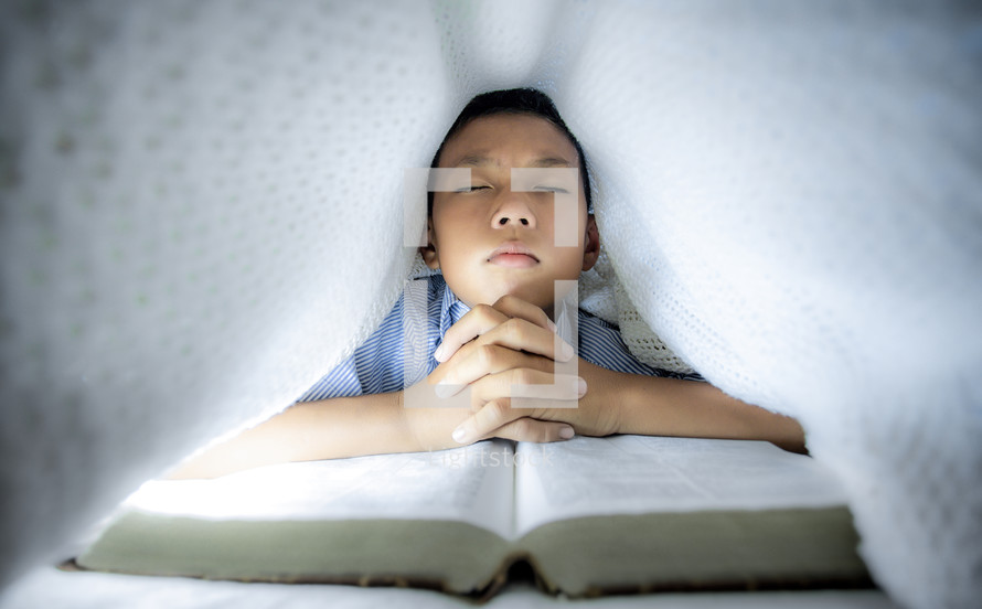 a little boy reading a Bible and praying under a blanket 