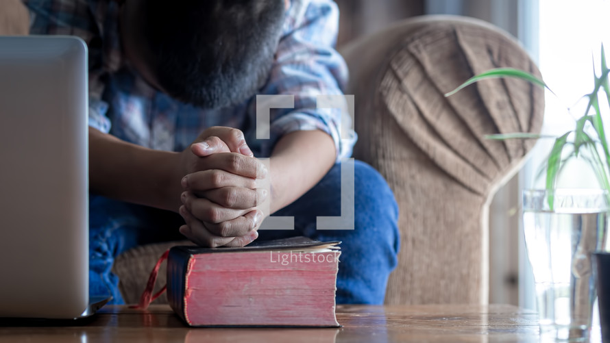 a boy praying over a Bible sitting in front of a laptop at home 