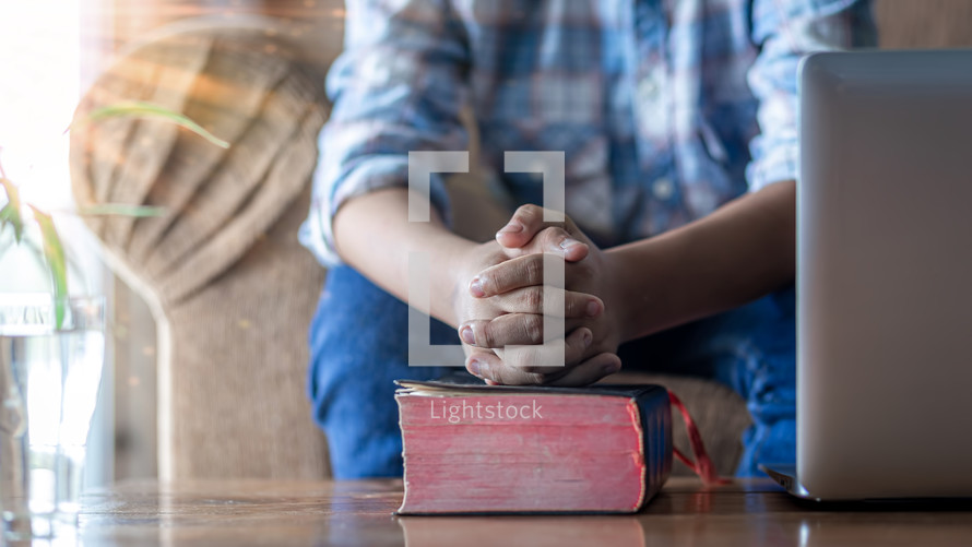 a boy praying over a Bible in front of a laptop at home 