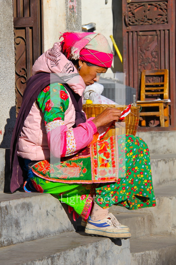 Woman in traditional Chinese minority tribe clothes sewing whilst sitting on stairs