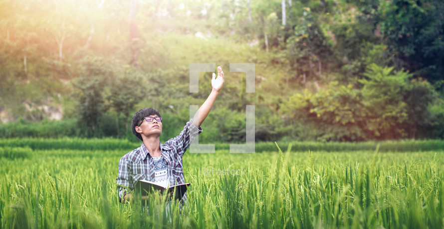 a young man holding a Bible with hand raised standing in a rice field 