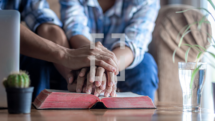 closeup of two people with hands on a Bible praying at home 