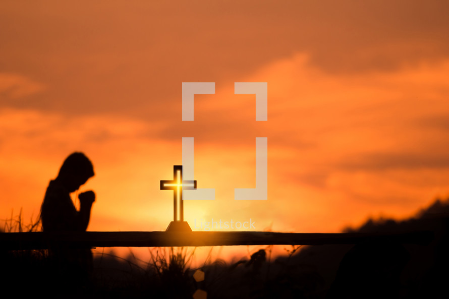 A boy praying to God with a cross at sunset 