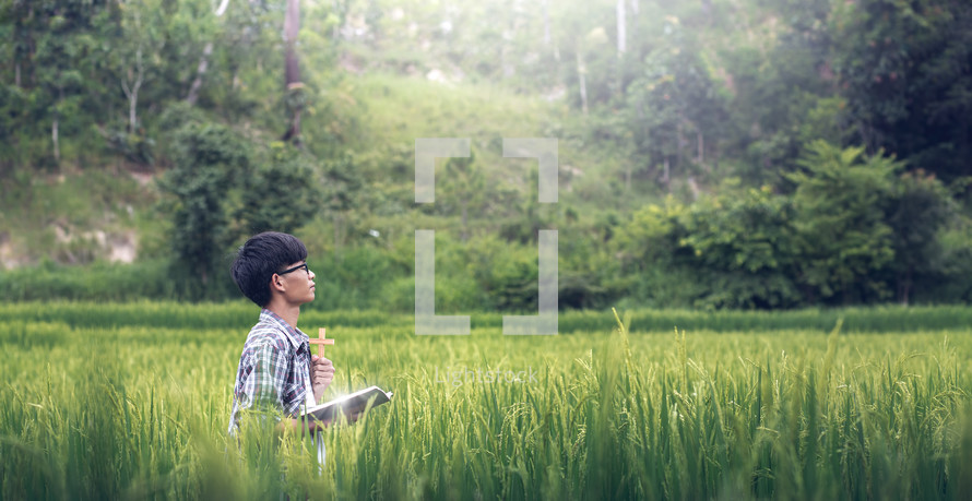 a young man holding a Bible praying in a rice field 