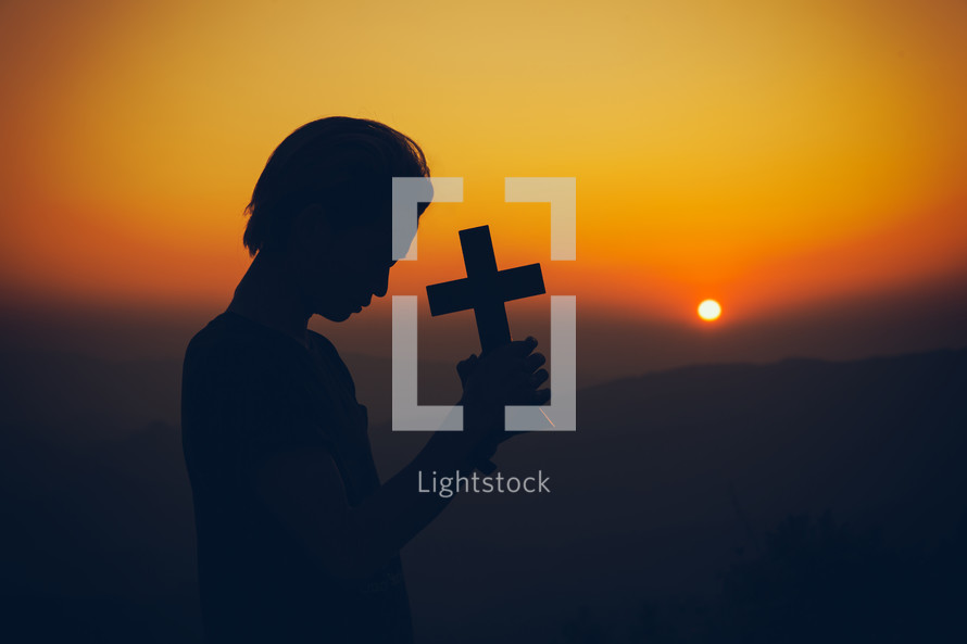 Young man praying and holding Cross at sunset 