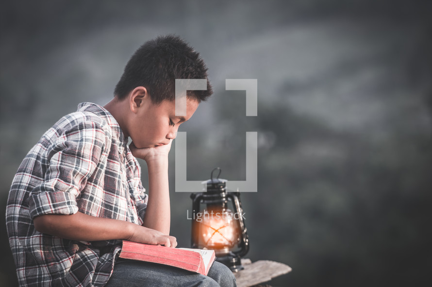 a boy reading a Bible outdoors by a lantern 