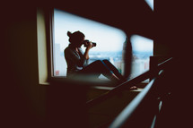 woman with a camera taking a picture sitting in a window sill 