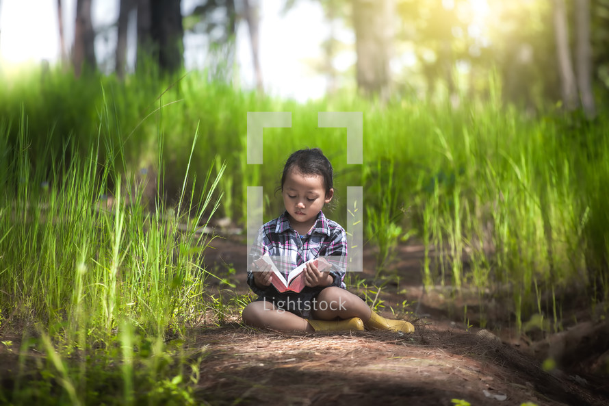 a little girl sitting outdoors in tall grass in a forest reading a Bible 