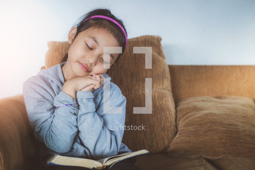 a girl reading a Bible and praying while sitting on a couch 