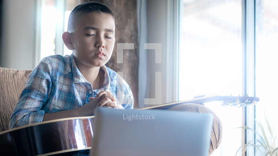 Boy with a guitar praying in front computer laptop, Online church in home, Home church during quarantine coronavirus Covid-19