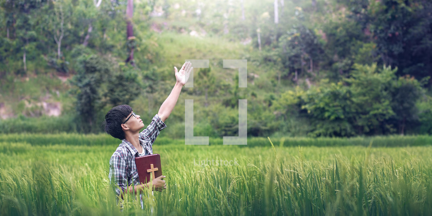a young man holding a Bible with hand raised standing in a rice field 