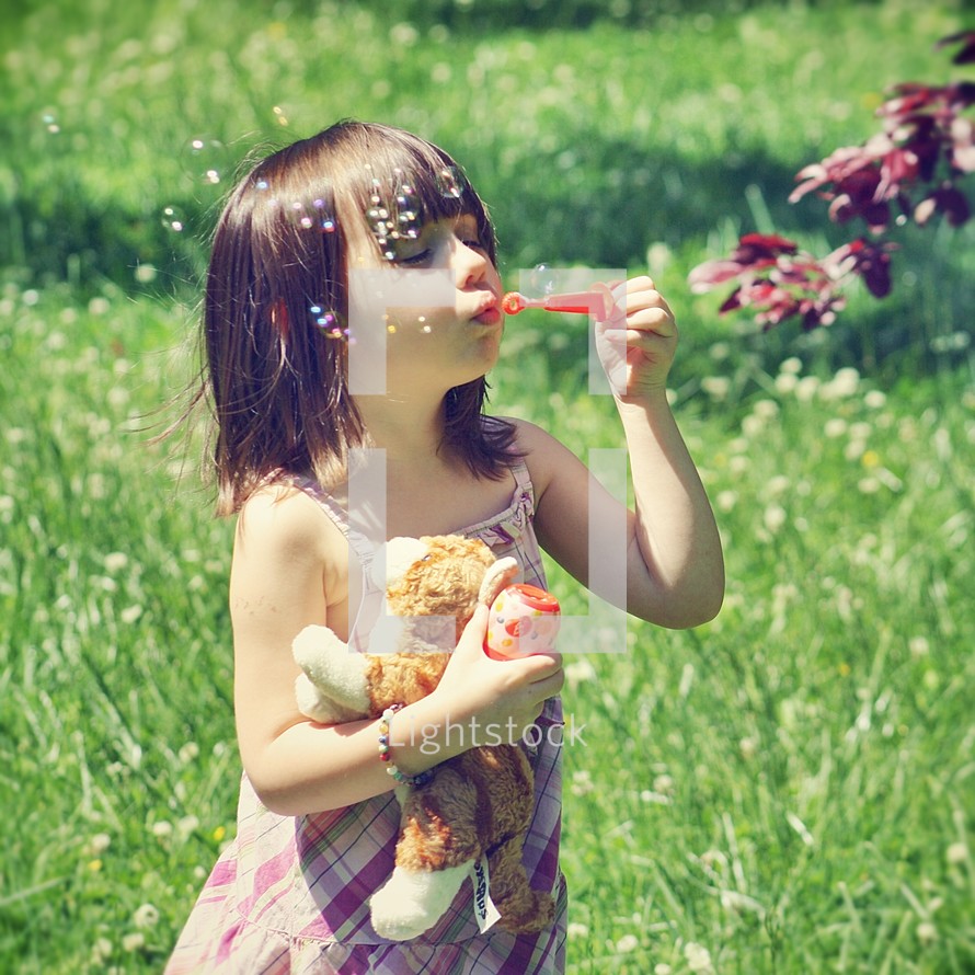 Girl holding stuffed animal while blowing bubbles outdoors.