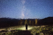 a man holding a flashlight standing under a starry sky surrounded by mountains 
