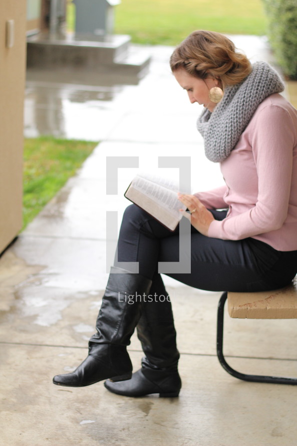 a woman sitting on a bench outdoors reading a Bible in the rain 