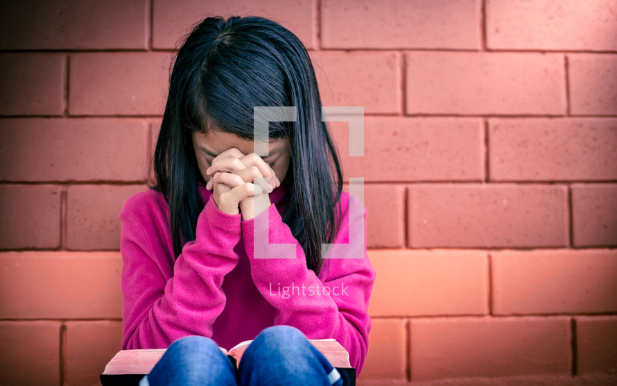 a girl child praying in front of a brick wall 