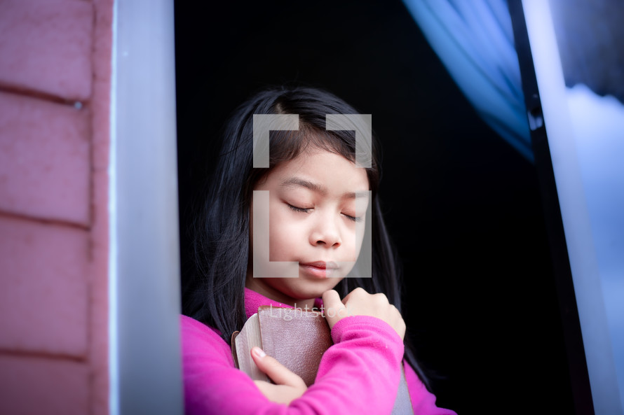 a girl child holding a Bible praying in an open window during quarantine 