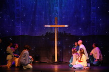 performers on stage kneeling in front of a cross and a scene of Bethlehem 