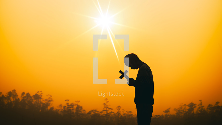 a male holding a cross at sunset against an orange sky