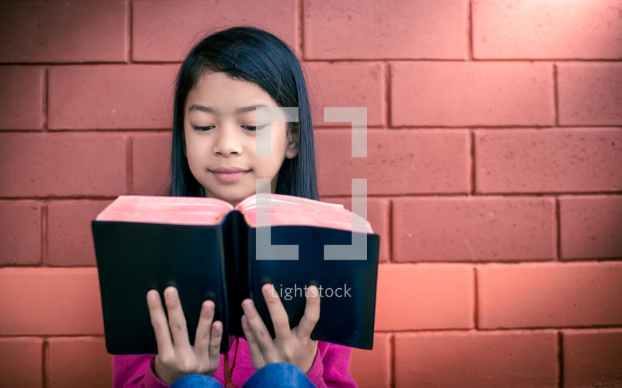 a girl reading a Bible outdoors in front of a brick wall 