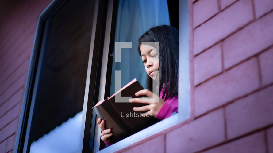 a girl child reading a Bible in an open window during quarantine 