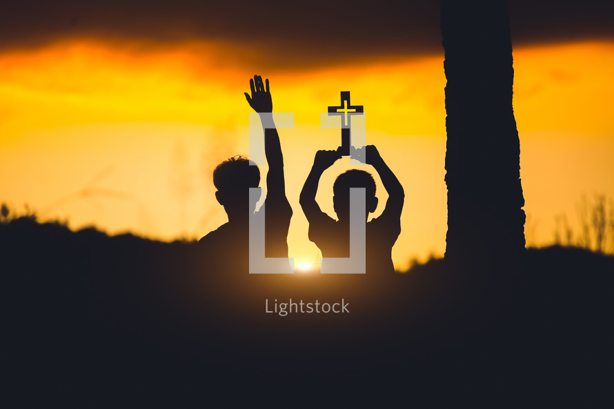 two boys with hands raised holding a cross against an orange sky at sunset 