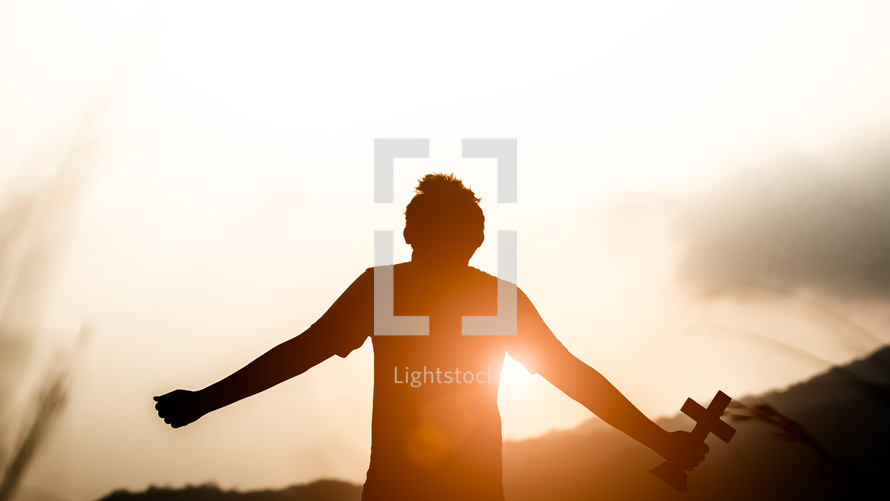 outstretched arms holding up a cross with sunlight at sunset in the background
