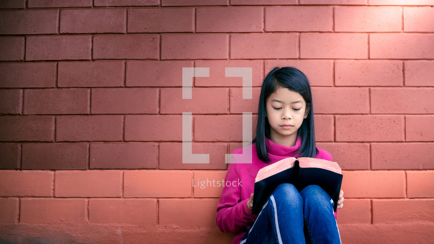 a girl reading a Bible in front of a brick wall 