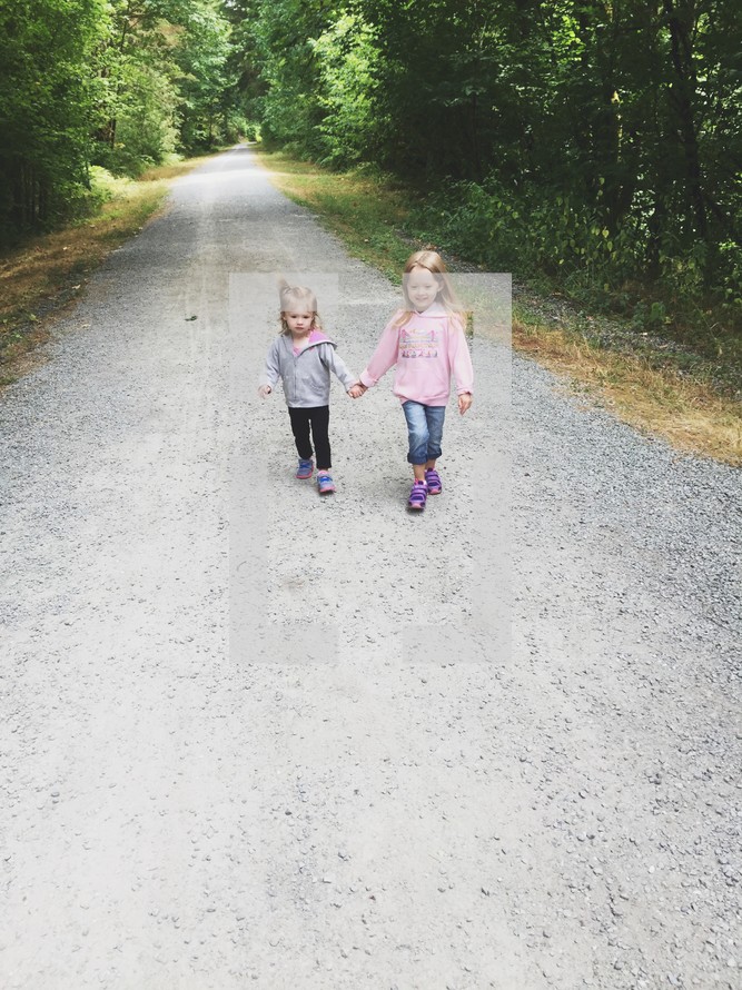 sisters holding hands walking on a gravel road 
