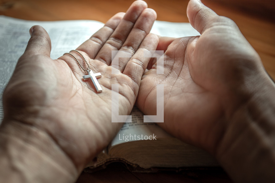 cross necklace in open hands in prayer over a Bible 