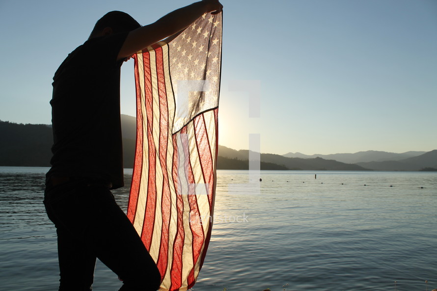 Sunlight behind a silhouette of a man holding and American flag over water.