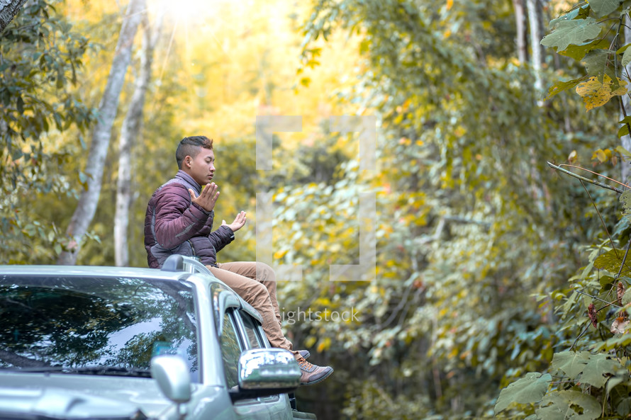 a young man sitting on top of his vehicle praying outdoors in a forest 