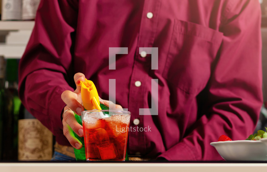 Close-up of a man's hands mixing a vibrant red cocktail at a bar with a soda siphon.