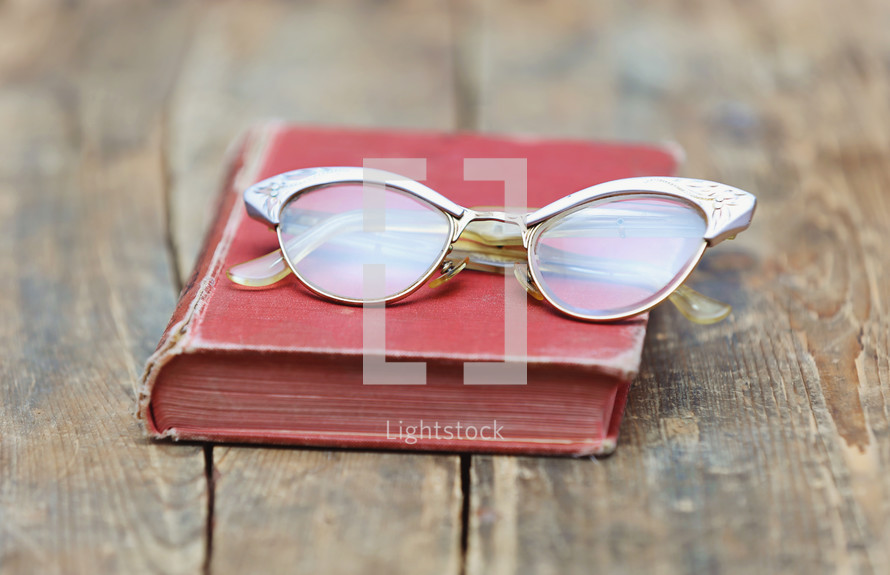 Eye glasses on an old red book, which is on a rugged wooden table.