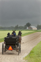 Horses pulling a carriage on a dirt road toward a farmhouse.