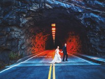A bride and groom walking across a street in front of a tunnel. 