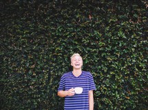 A man standing in front of an ivy covered wall drinking coffee. 