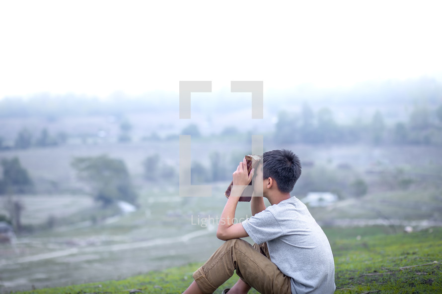 a boy sitting on a hill praying holding a Bible 